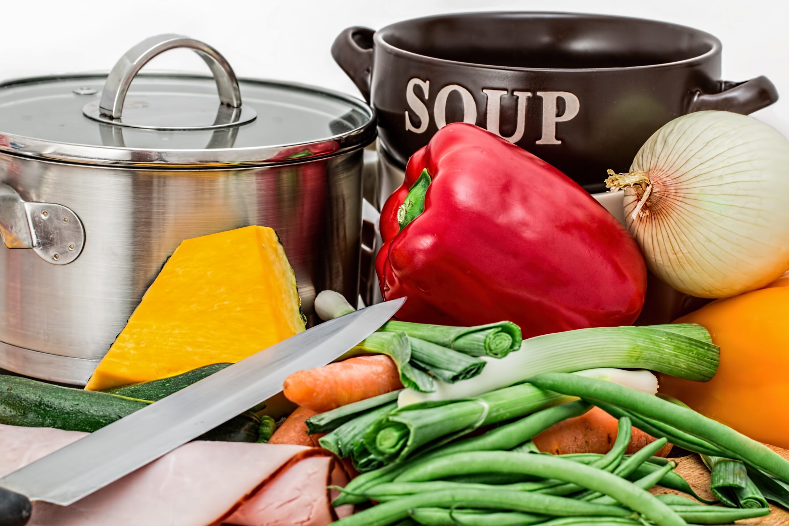 Assorted vegetables including green beans, peppers, and onions, with a soup pot, and soup bowl with the word Soup written on it and a knife nearby. Image source Pixabay